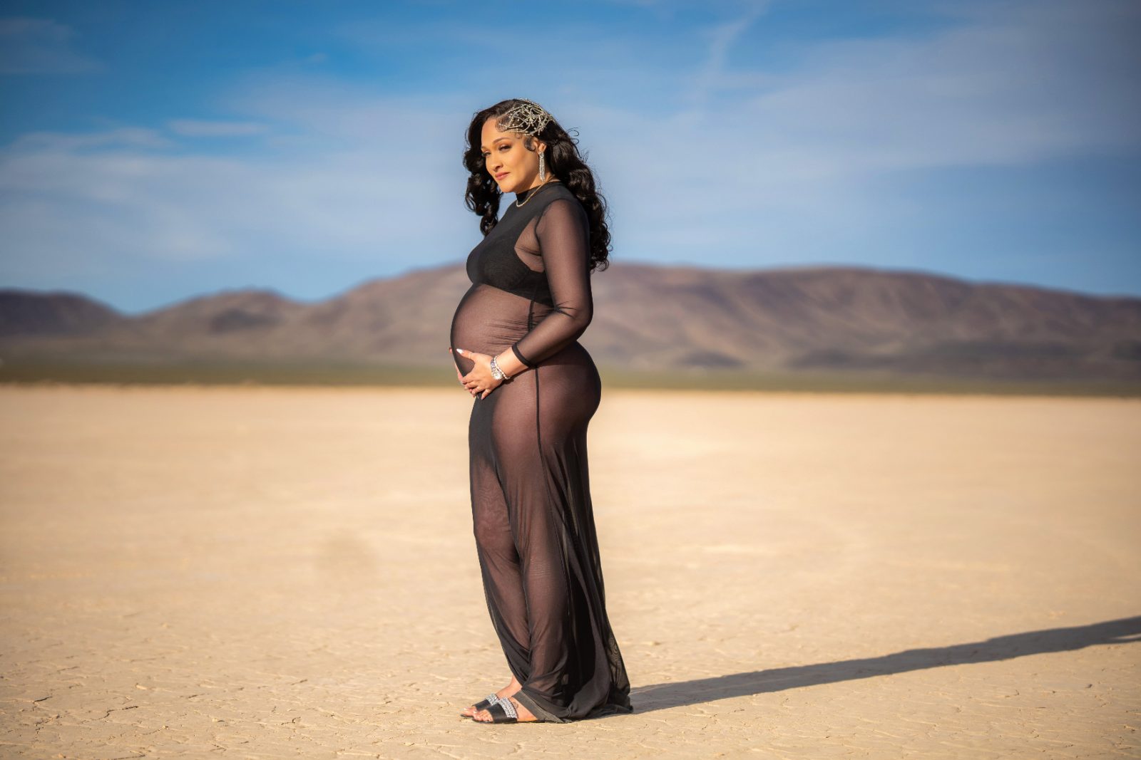 Las Vegas maternity photographer — expectant mother in a sheer black gown on a dry lake bed with desert mountains behind her